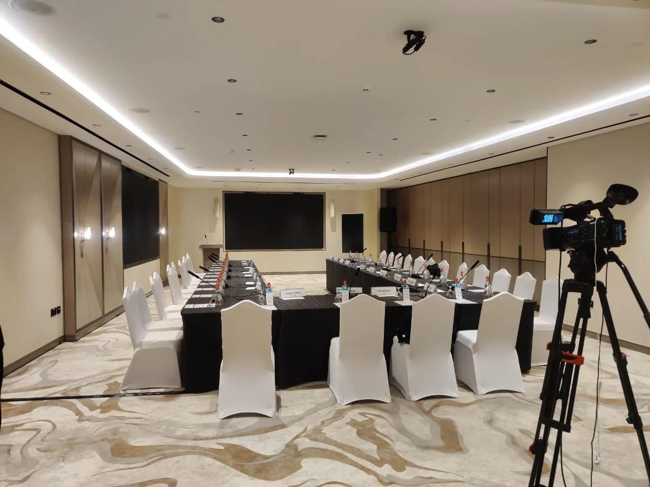 Broadcast camera positioned beside a U-shaped conference meeting setup.