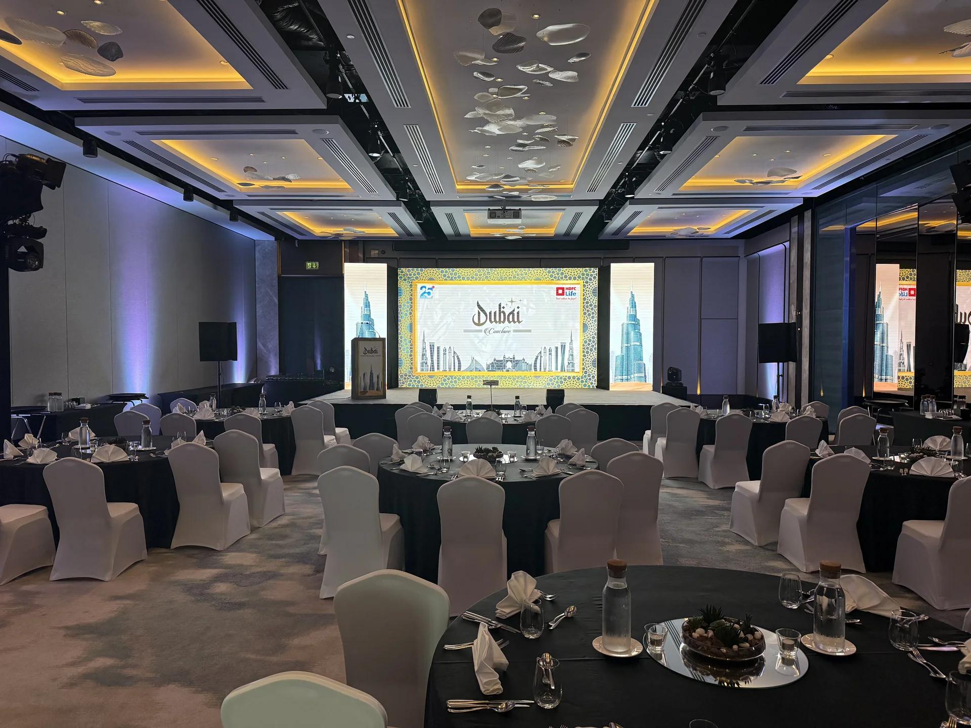 Conference banquet room with round tables and a multi-screen stage.